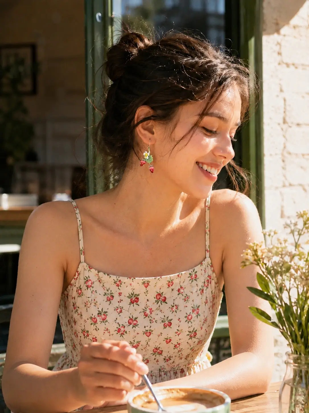 Woman wearing strawberry blossom vine drop earrings while seated at a sunlit outdoor café.