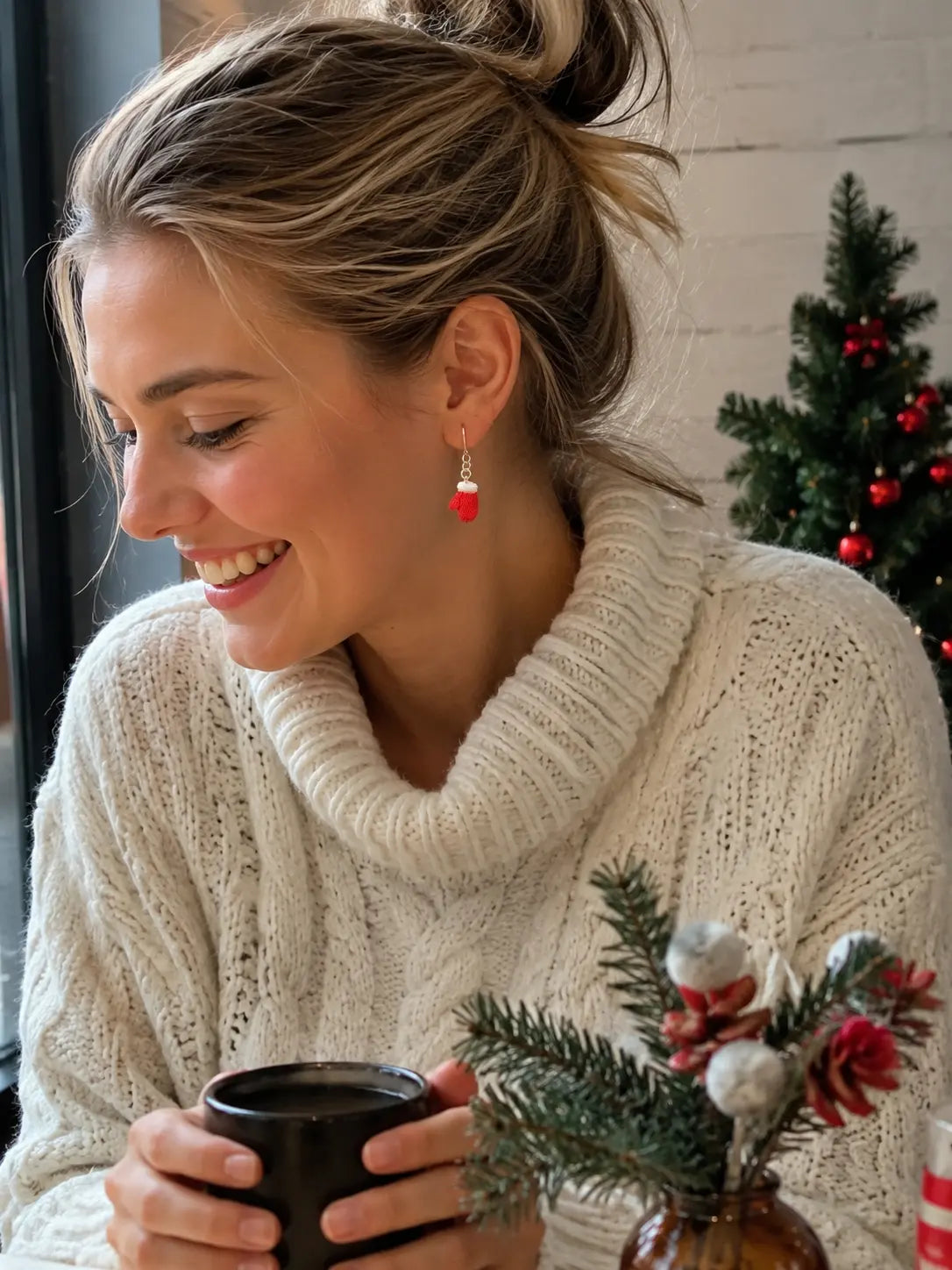 Woman wearing festive Santa mitten crochet earrings while holding a warm drink beside Christmas decorations