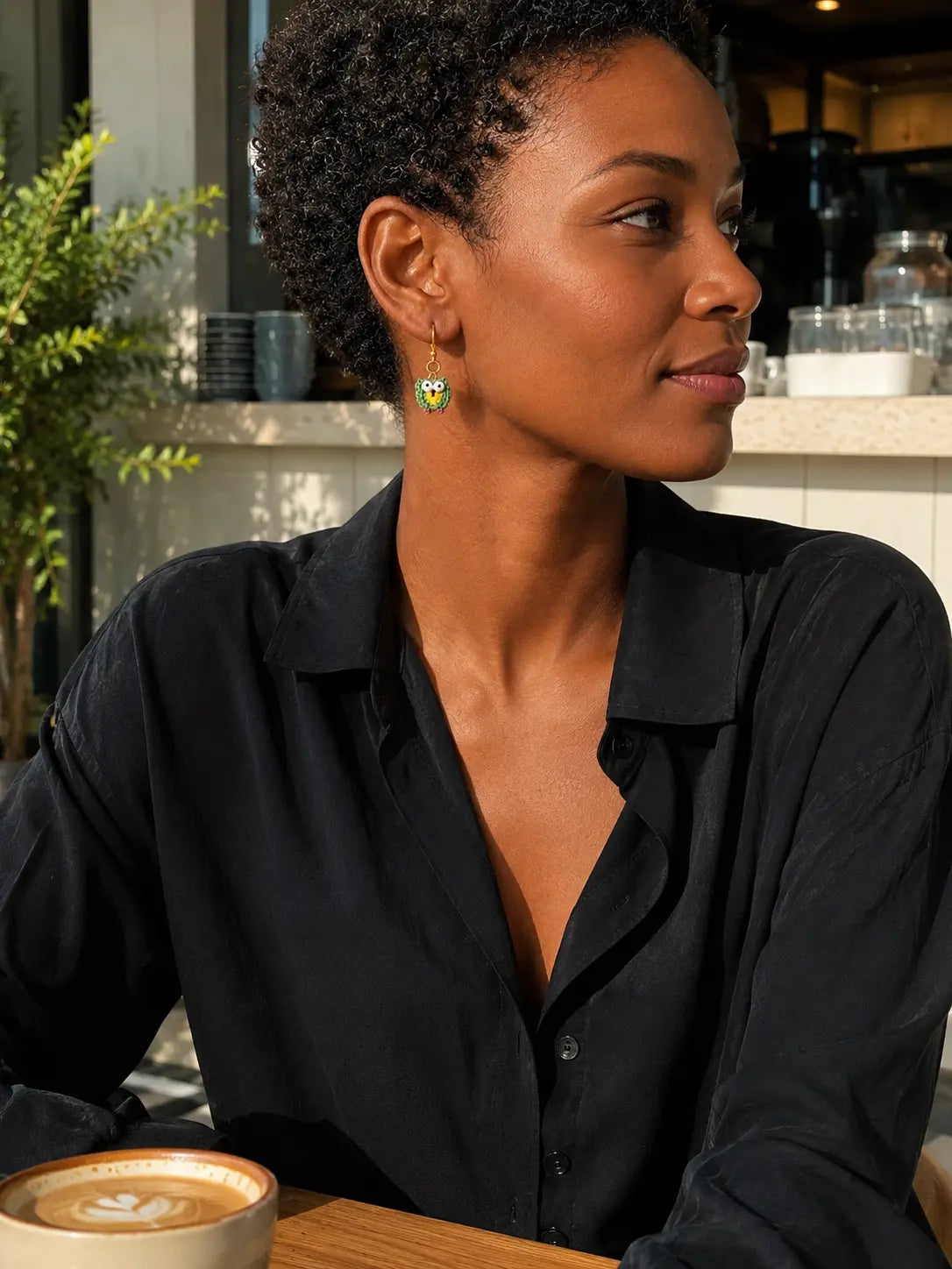 Side profile of a woman wearing green micro crochet owl drop earrings while seated in a softly lit café.