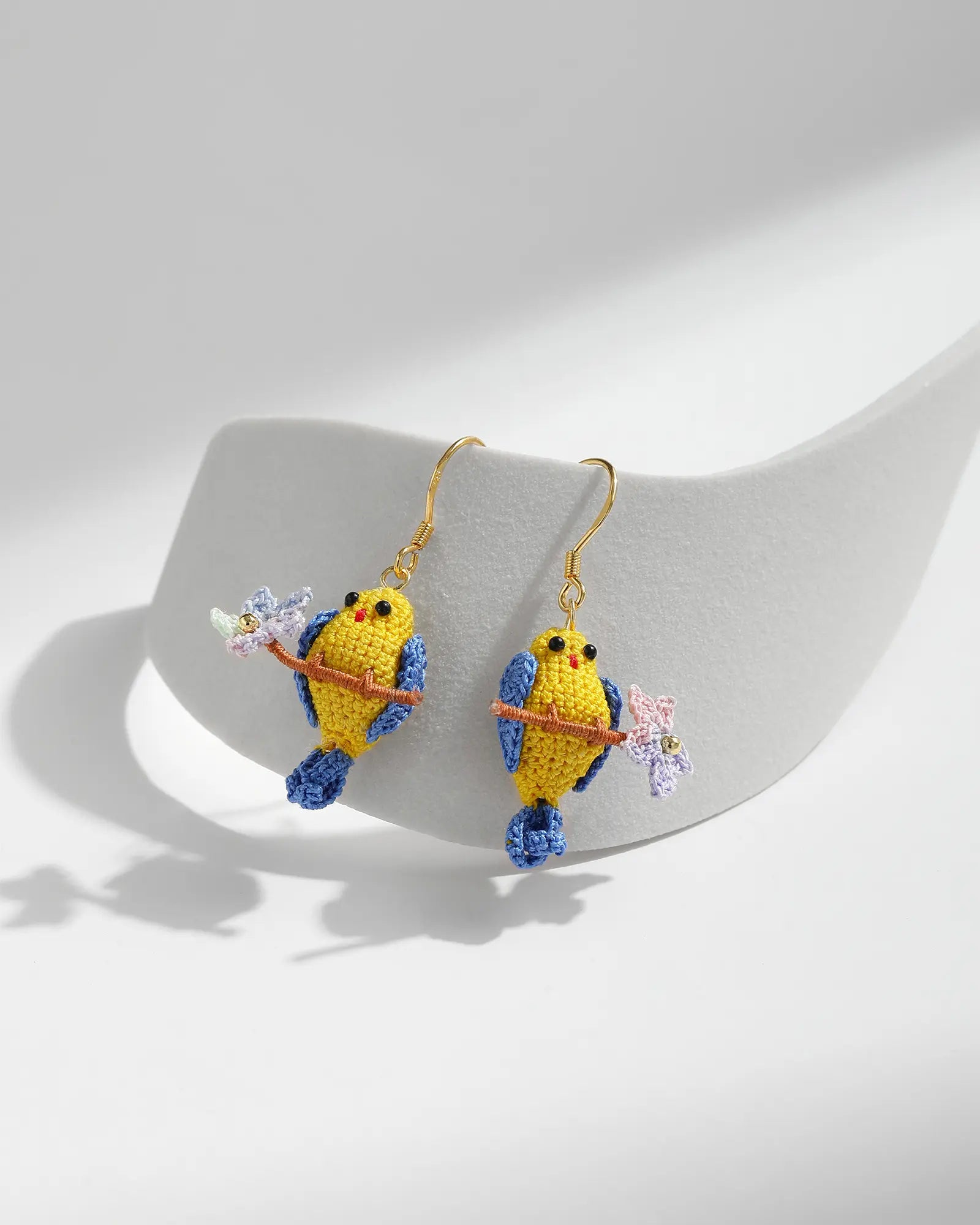 Yellow micro crochet parrot and blossom branch drop earrings with blue tails and gold hooks on a white jewellery display.