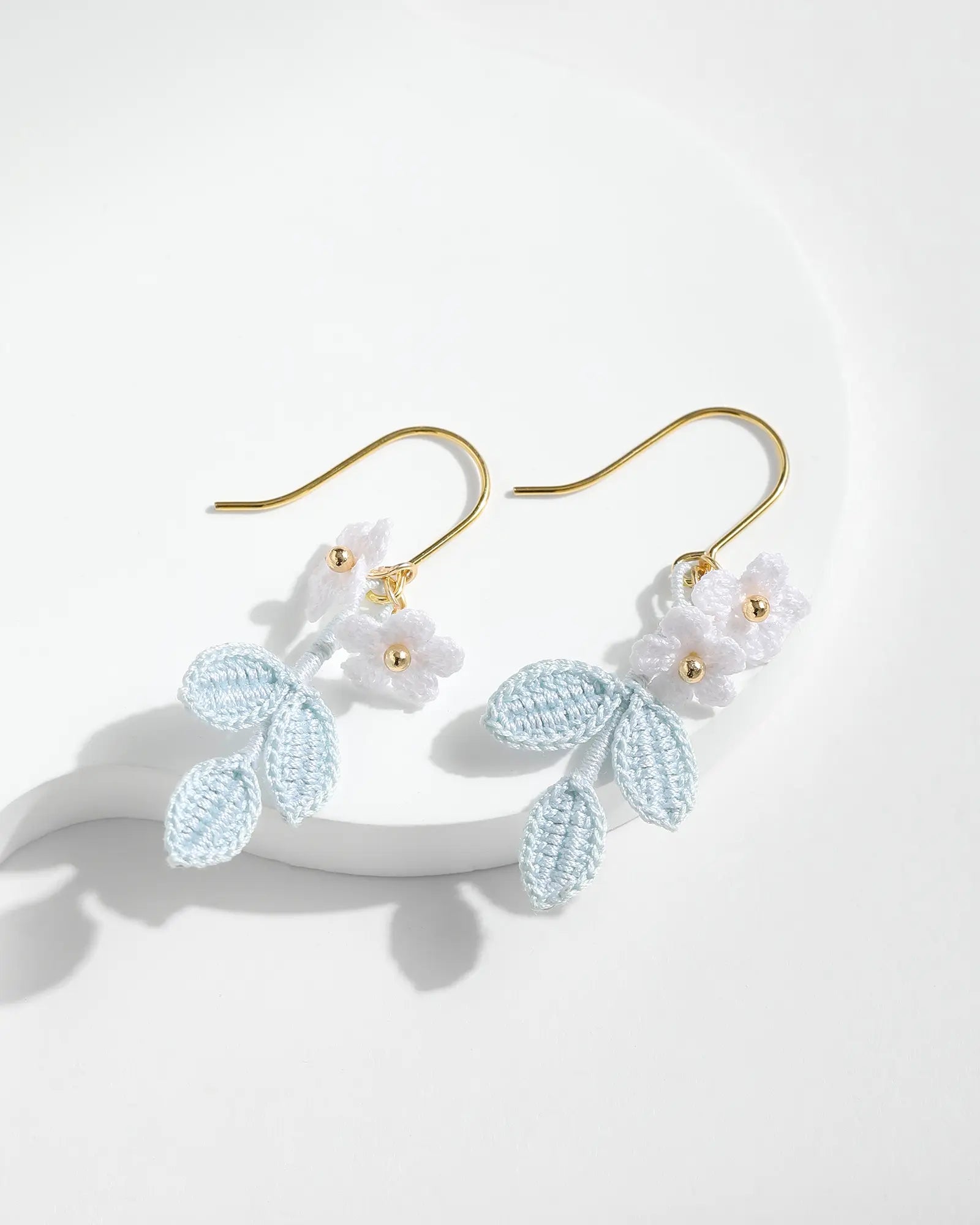 Pair of powder blue leaf and white blossom crochet drop earrings with gold-tone hooks displayed on a white background.
