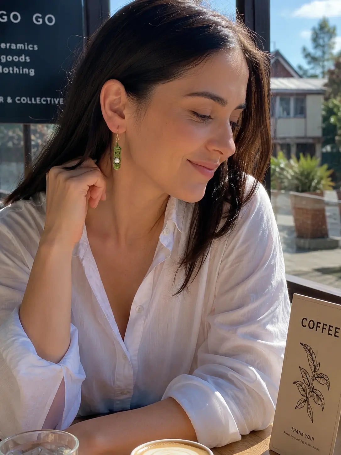 Model wearing micro crochet Pea Pod Earrings at a sunny café, showing the green dangle earrings from the side.