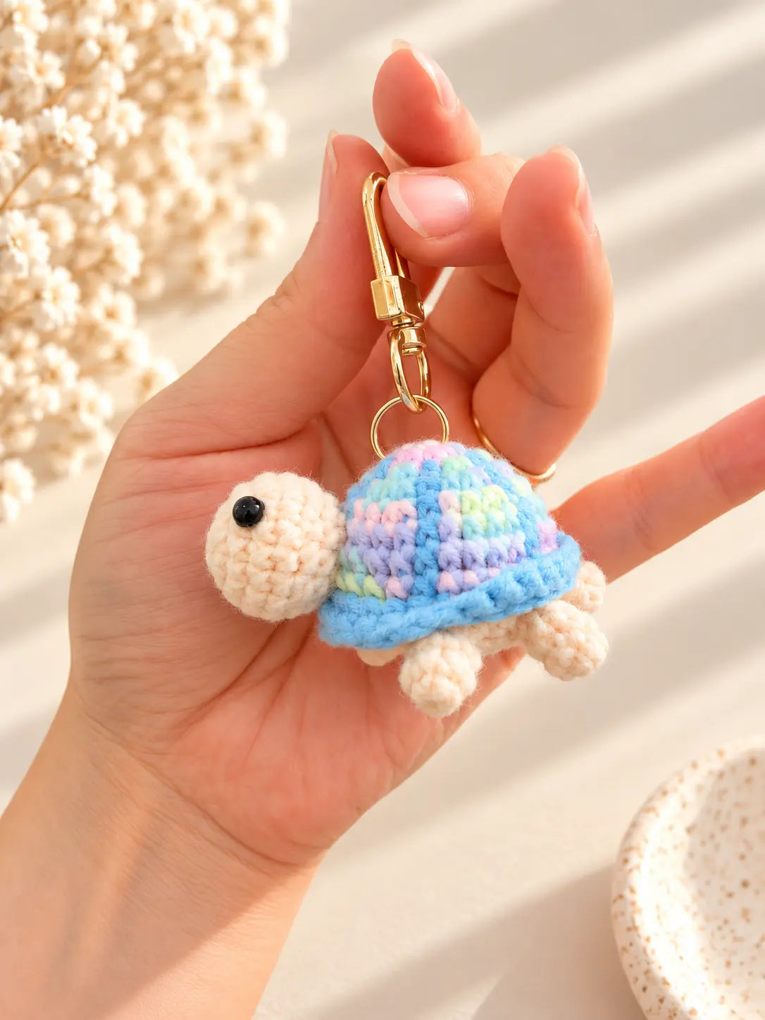 Hand holding a cute handmade crochet turtle keychain with a pastel blue and purple shell, cream-coloured head, tiny legs and gold metal clasp.