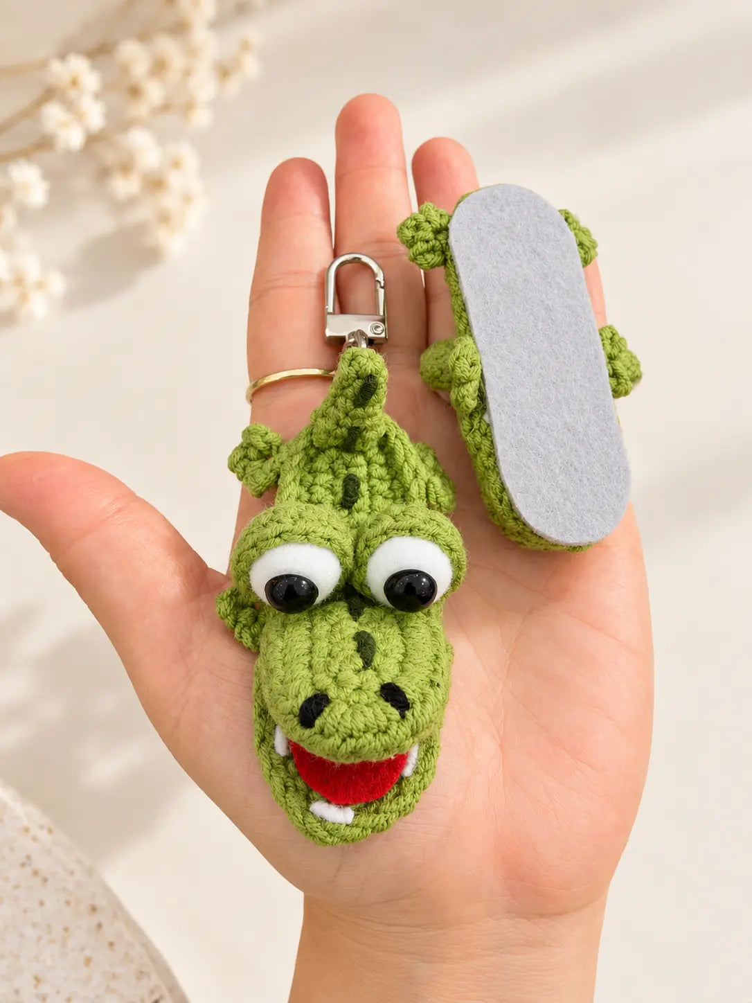 Hand holding a cute handmade flat crochet crocodile keychain with googly eyes, a bright red mouth and green textured stitching, showing its size and details.