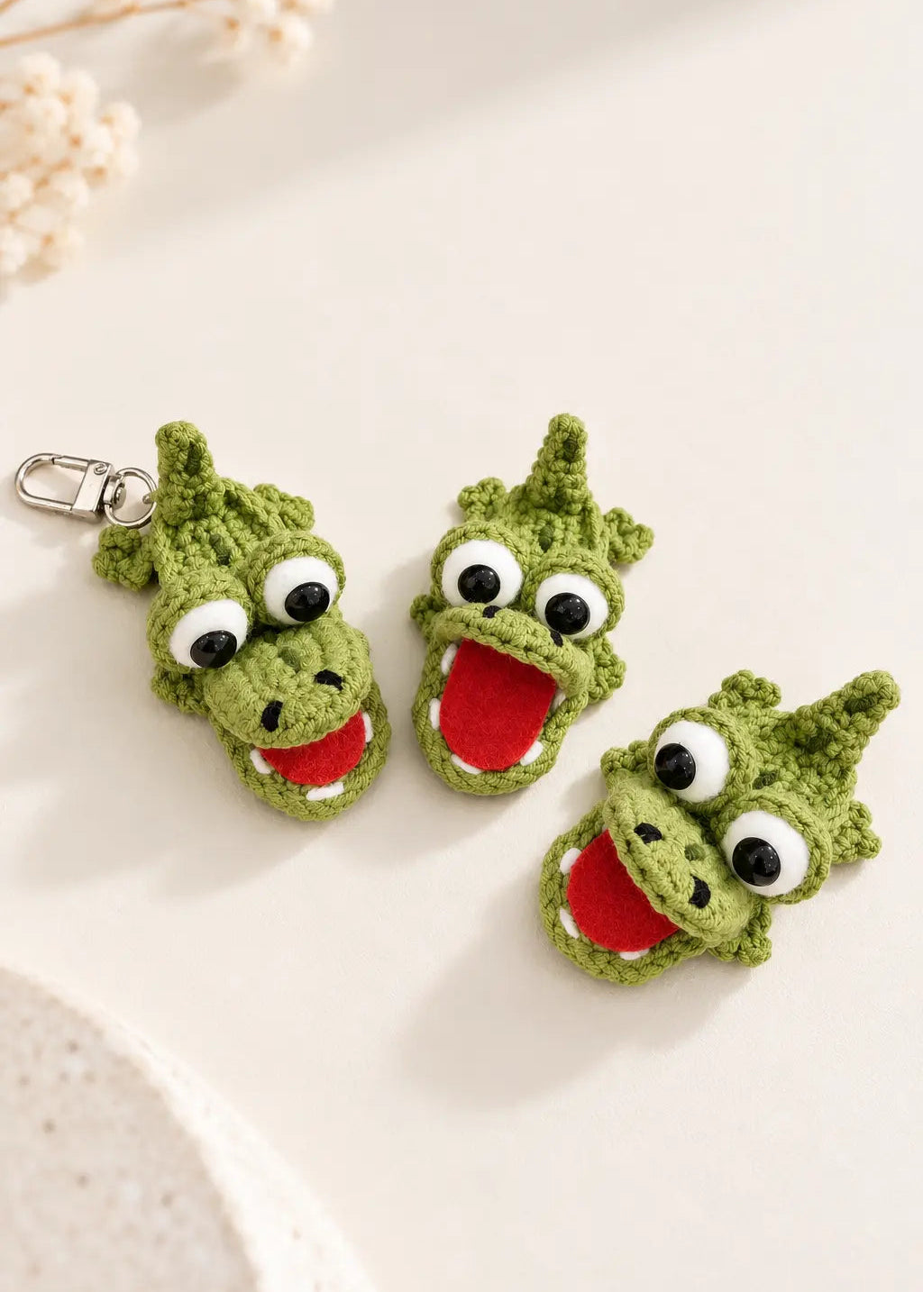 Three cute green flat crochet crocodile keychains with playful googly eyes and open red mouths, arranged on a soft cream background.