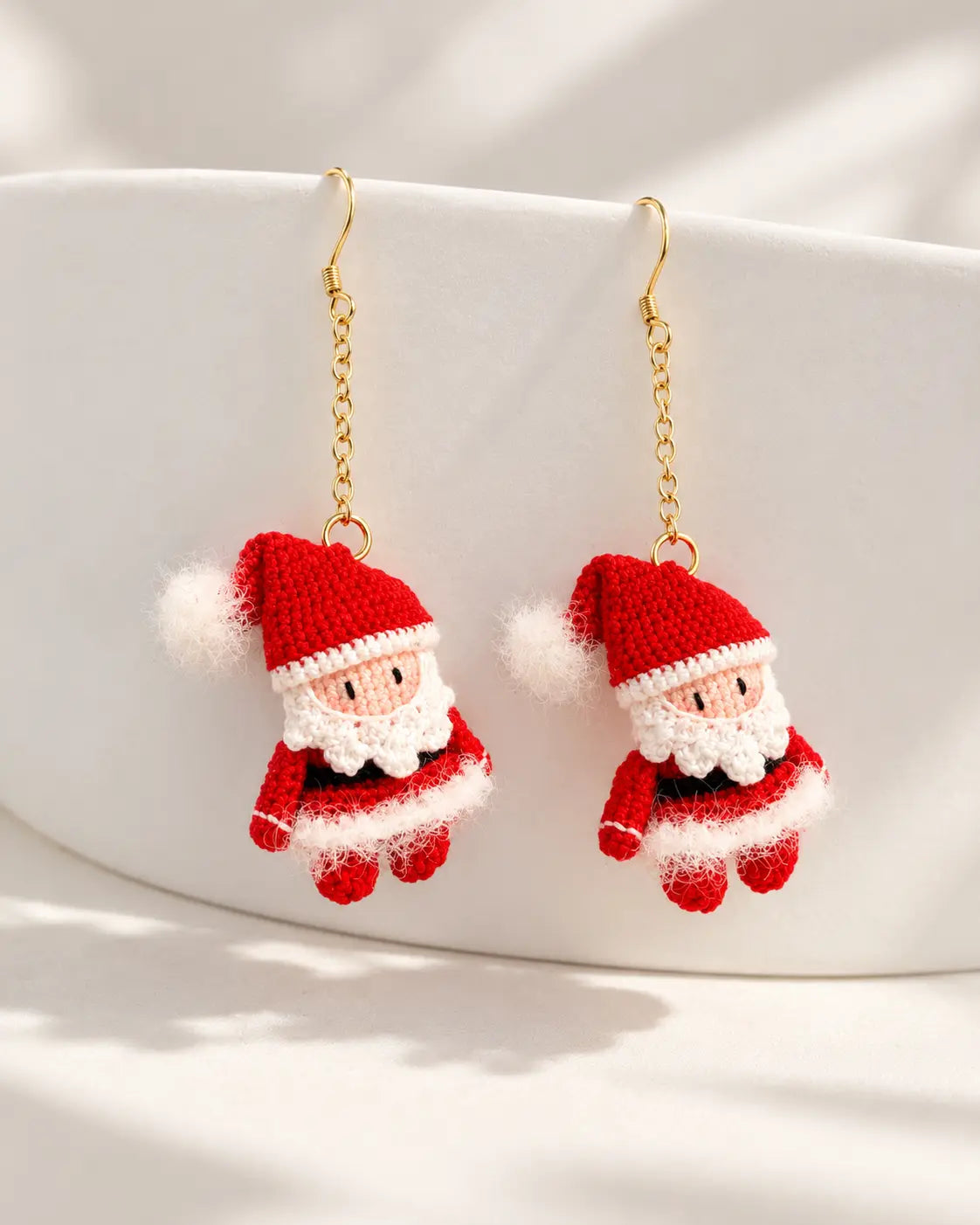 Pair of miniature crochet Santa Claus drop earrings with red hats, white beards and gold-tone hooks against a minimalist cream background.