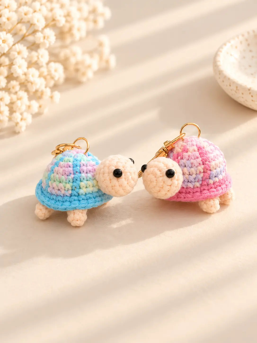Two adorable handmade crochet turtle keychains with pastel pink and blue shells, cream-coloured heads and tiny legs, displayed together on a soft beige background.