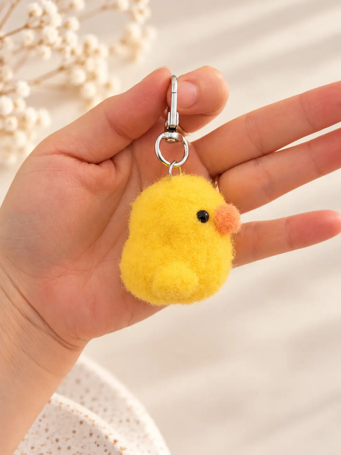 Close-up of a hand holding a fluffy yellow crochet duck keychain with a soft fuzzy texture, tiny orange beak and silver metal keyring.