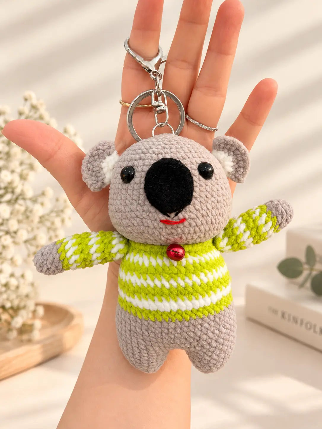 Handmade large crochet koala keychain wearing a green striped sweater and red bell collar, displayed on a hand.