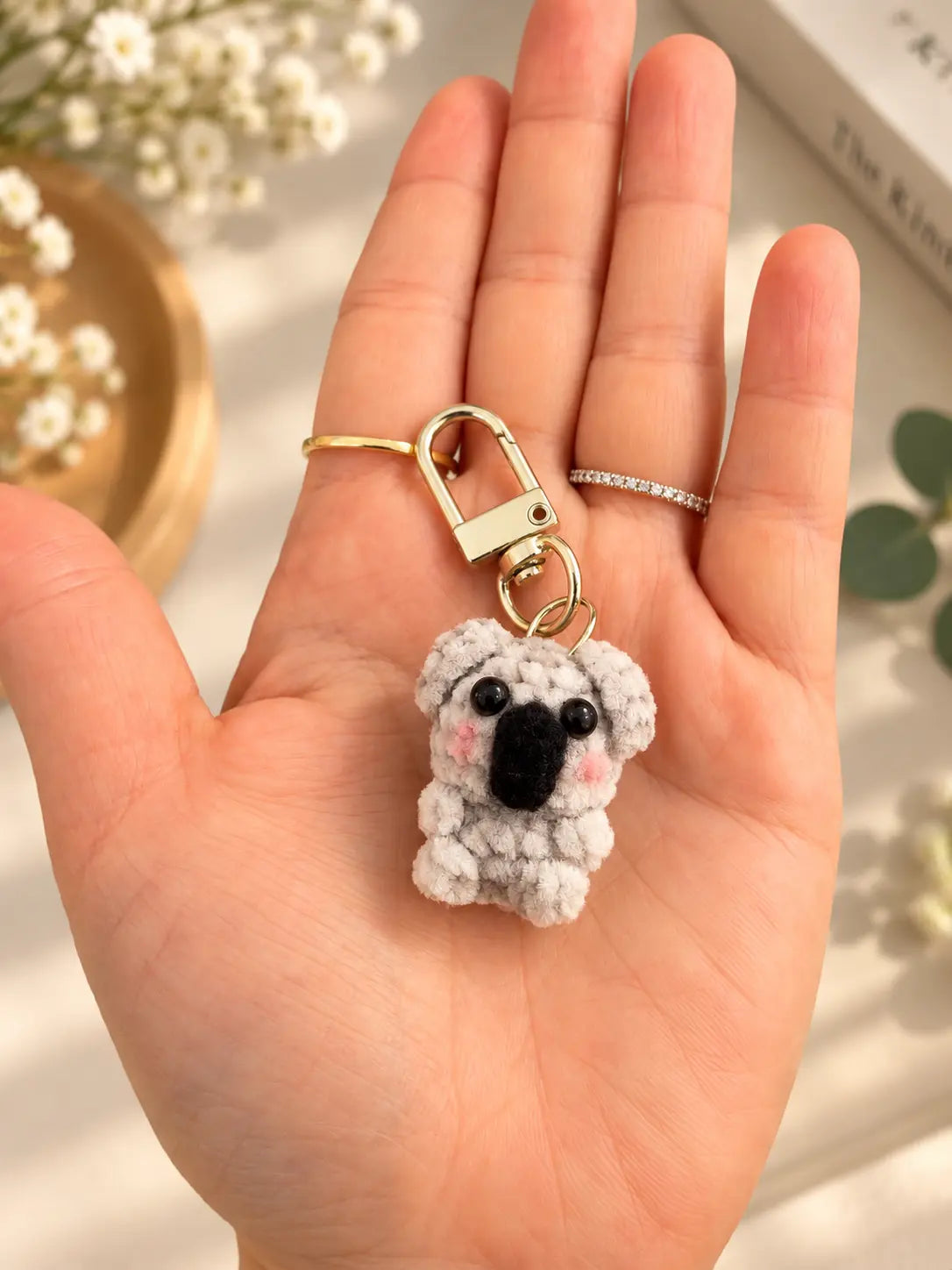 Handmade mini crochet koala keychain displayed on a hand, featuring a tiny grey koala with black nose and white belly.
