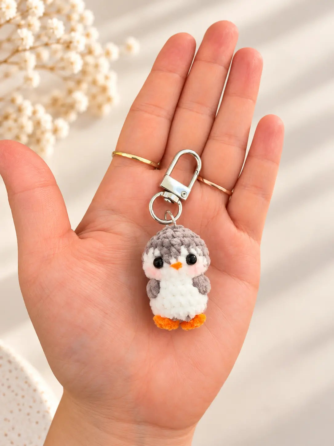 Hand holding a tiny handmade grey and white crochet penguin keychain with a yellow beak, orange feet and silver metal clasp, showing its miniature size.