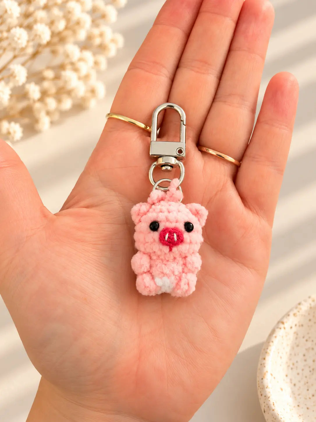 Hand holding a tiny handmade pink crochet pig keychain with a round snout, black eyes, little ears and a silver metal clasp, showing its miniature size.