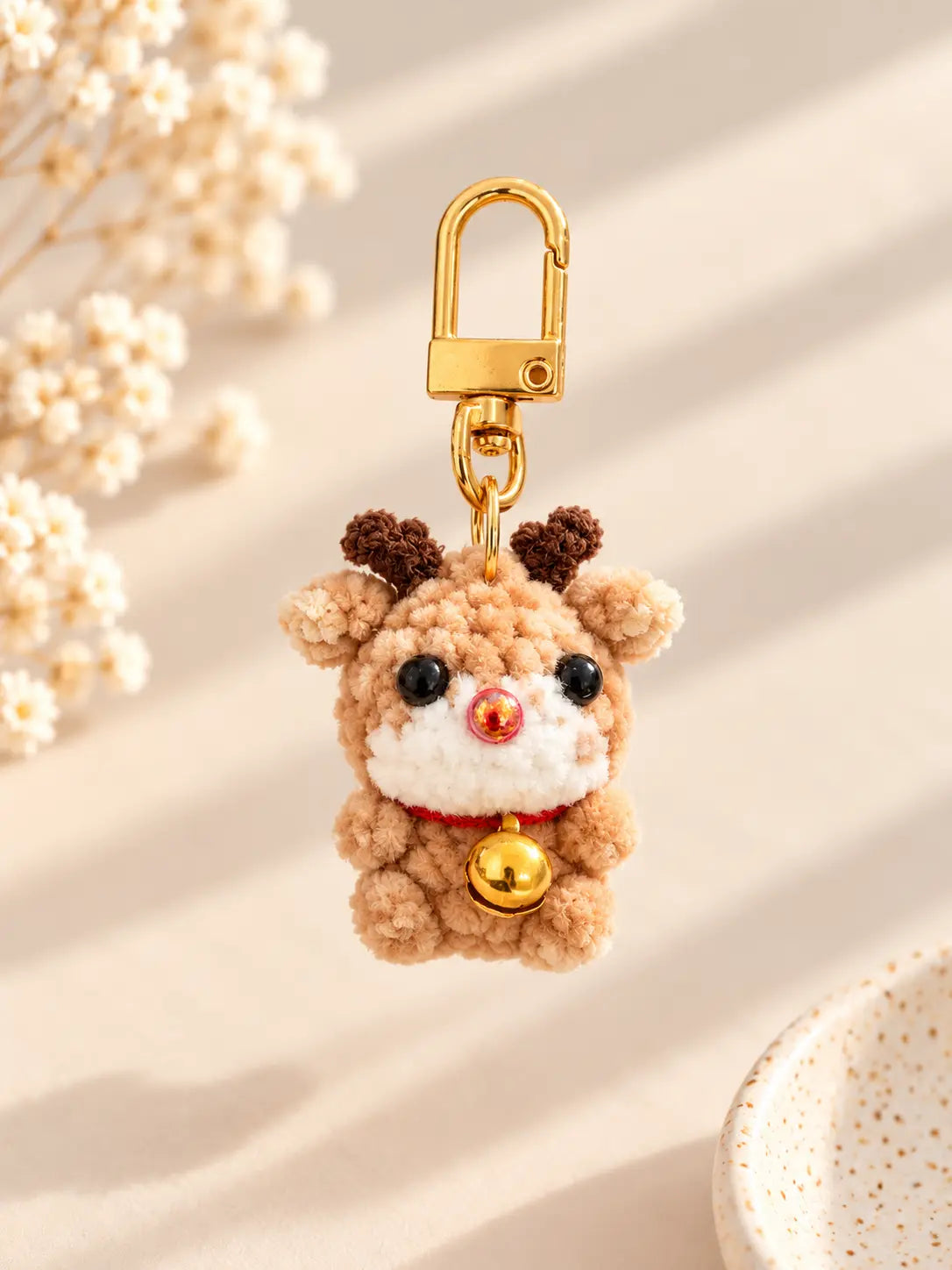 Adorable handmade mini crochet reindeer keychain featuring brown antlers, a red nose, a tiny gold bell and gold metal clasp, displayed against a soft cream background.