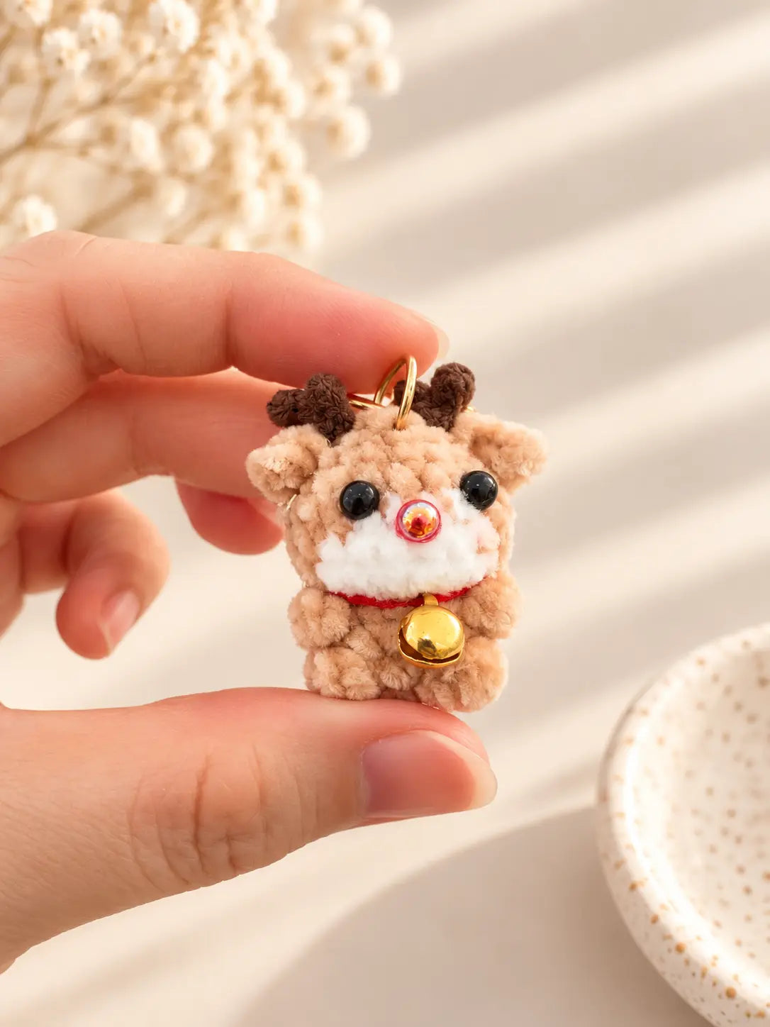 Hand holding a cute handmade mini crochet reindeer keychain with brown antlers, a red nose, white muzzle and tiny gold bell, showing its miniature size.