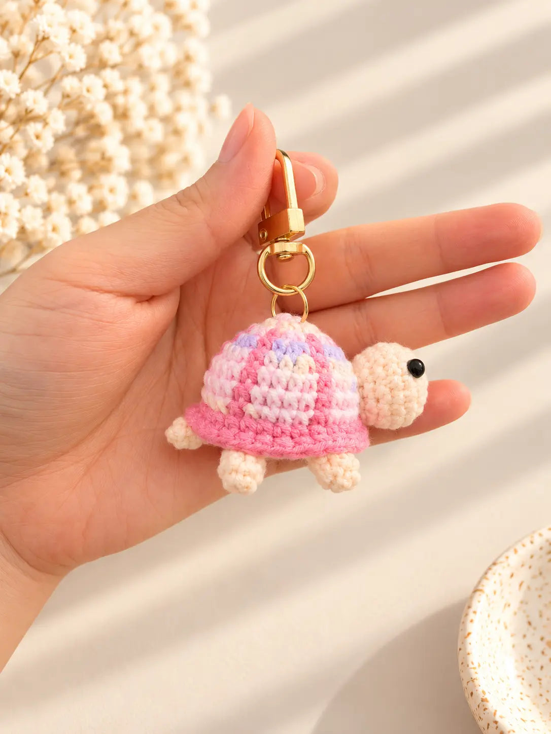 Hand holding a cute handmade crochet turtle keychain with a pastel pink shell, cream-coloured head and tiny legs, featuring a gold metal clasp.