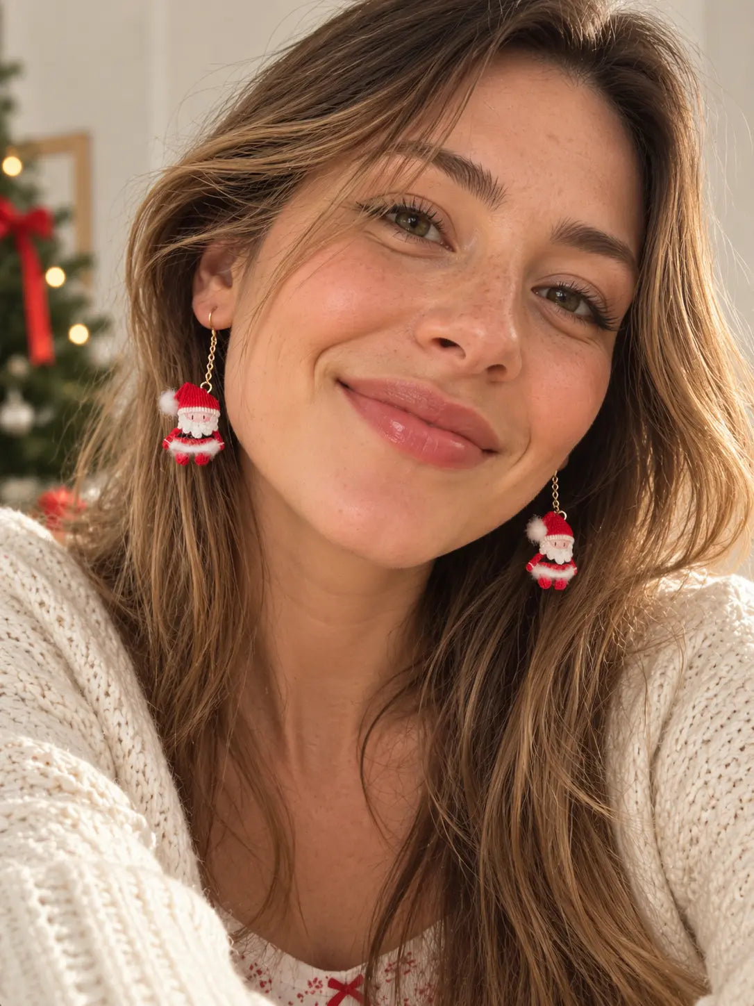 Smiling woman wearing a pair of miniature crochet Santa Claus drop earrings with a cream knit sweater in a festive Christmas setting.