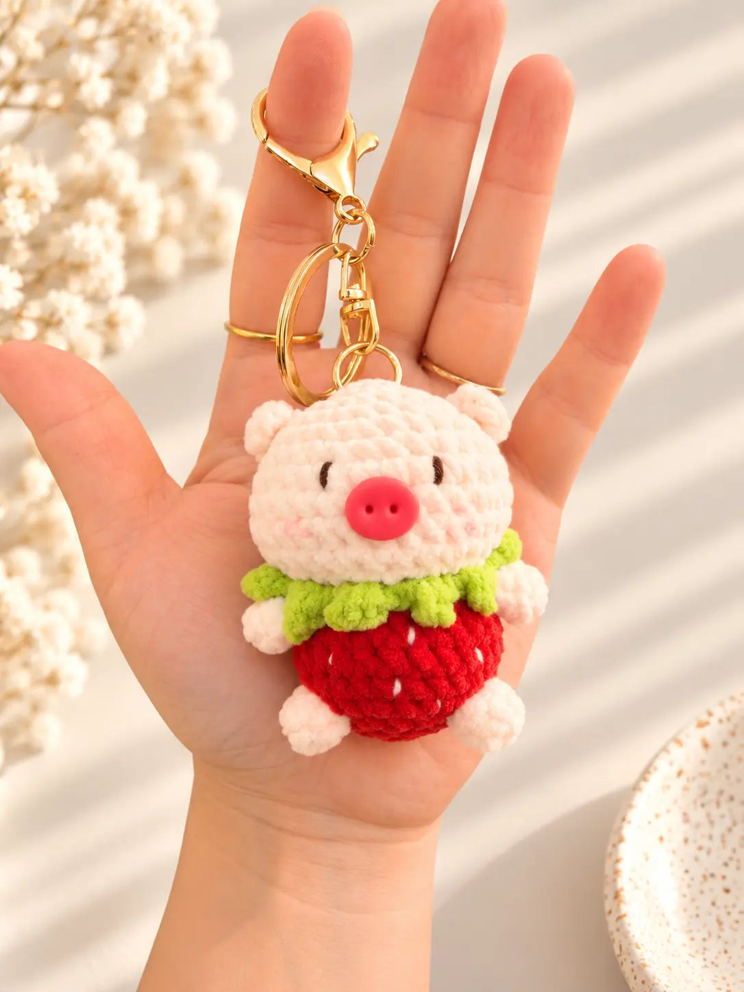 Hand holding an adorable handmade crochet pig keychain featuring a pink pig with a round snout, green leafy collar, red strawberry outfit and gold metal clasp.